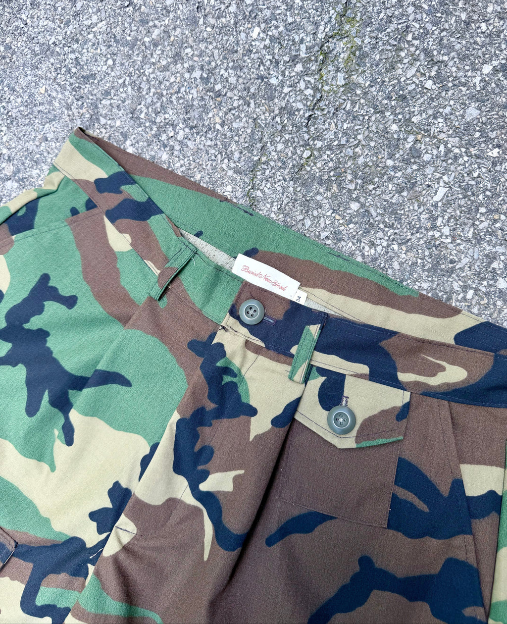 Close-up of camouflage cargo shorts laid on gray asphalt, showing the waistband, belt loops, button closure, and front pocket.